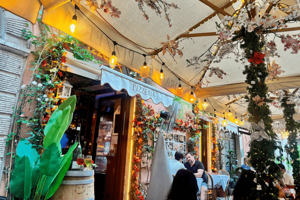 Terrazza esterna di Osteria della Vite a Roma, decorata con fiori e luci calde, con clienti seduti ai tavoli.